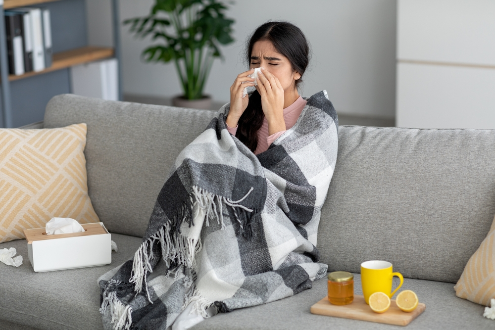 sick young woman blowing nose while wrapped in blanket on couch