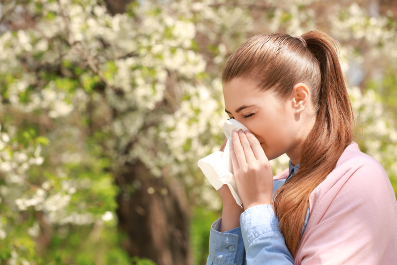 woman with seasonal allergies sneezing into tissue while walking past blooming tree